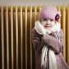 Furnace Maintenance Girl in winter clothes is shivering in front of radiator. Fearful expression. Concept for energy crisis or old heating system.
